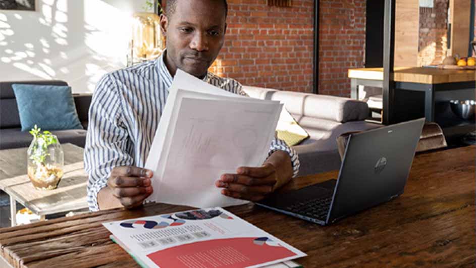 Een man zit aan een houten tafel met papieren, een laptop, en een document. Op de achtergrond is een bakstenen muur en een bank.