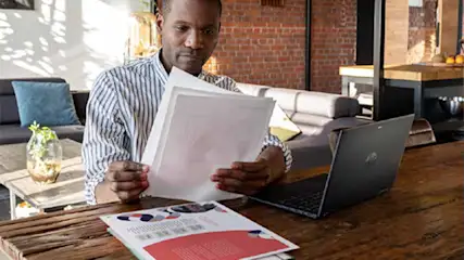 Een man zit aan een houten tafel met papieren, een laptop, en een document. Op de achtergrond is een bakstenen muur en een bank.