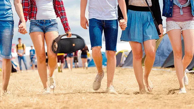 Five people walk on sand, holding hands. One carries a black speaker. Tents are in the background.