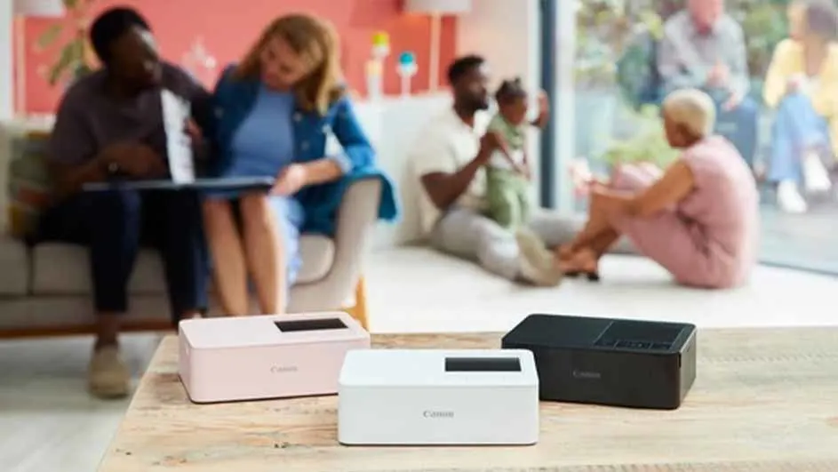 Drie Canon printers in roze, wit en zwart op een houten tafel. Op de achtergrond zitten mensen op een bank en op de vloer.