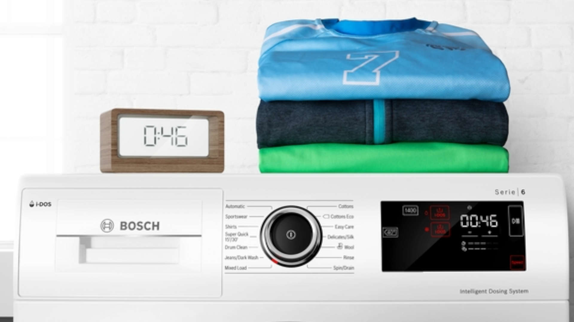 Wasmachine met digitale klok en stapel kleding erop. De klok geeft 0:46 aan. Bovenop liggen een blauw, grijs en groen shirt.