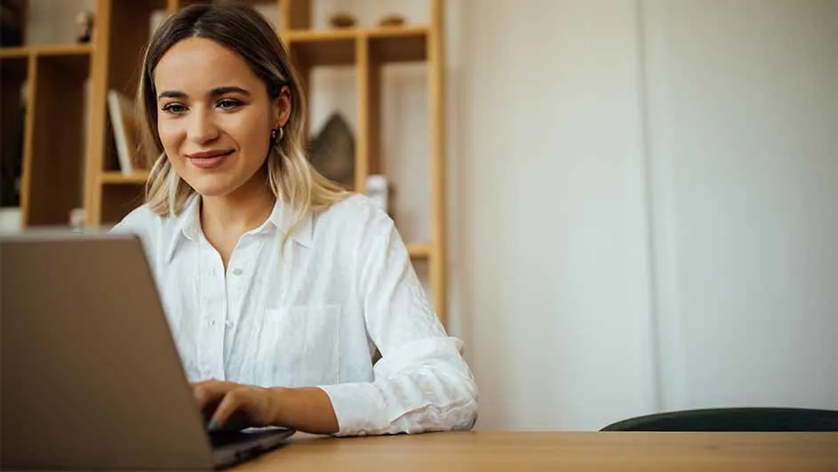 Een vrouw in een wit overhemd werkt achter een laptop op een houten bureau, met een boekenkast op de achtergrond.