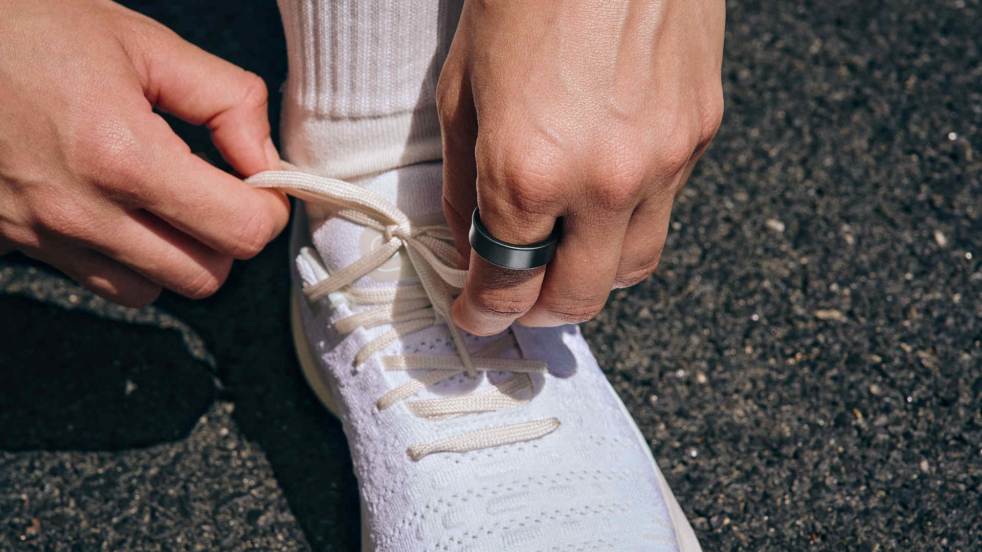 Twee handen strikken een witte schoenveter van een witte sportschoen. Een hand draagt een ring. Op de achtergrond ligt een donker oppervlak.
