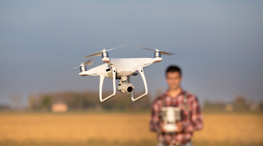 Een witte drone met camera zweeft in de lucht boven een bruin veld, met een vage figuur die een controller vasthoudt.