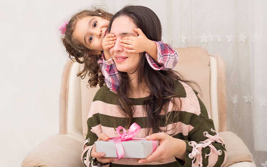 Klein meisje verrast haar moeder op de stoel, bedekt haar ogen terwijl mama een cadeautje met roze lint vasthoudt.