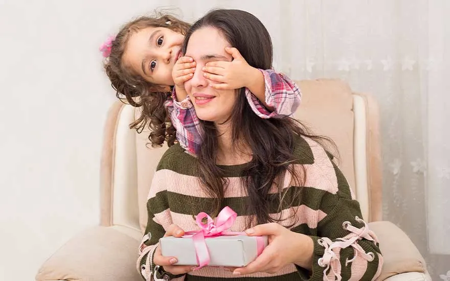 Klein meisje verrast haar moeder op de stoel, bedekt haar ogen terwijl mama een cadeautje met roze lint vasthoudt.