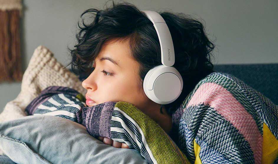 Iemand ligt ontspannen op een bank met een kleurrijke trui aan en draagt witte Sony koptelefoon, omringd door kussens en dekens.