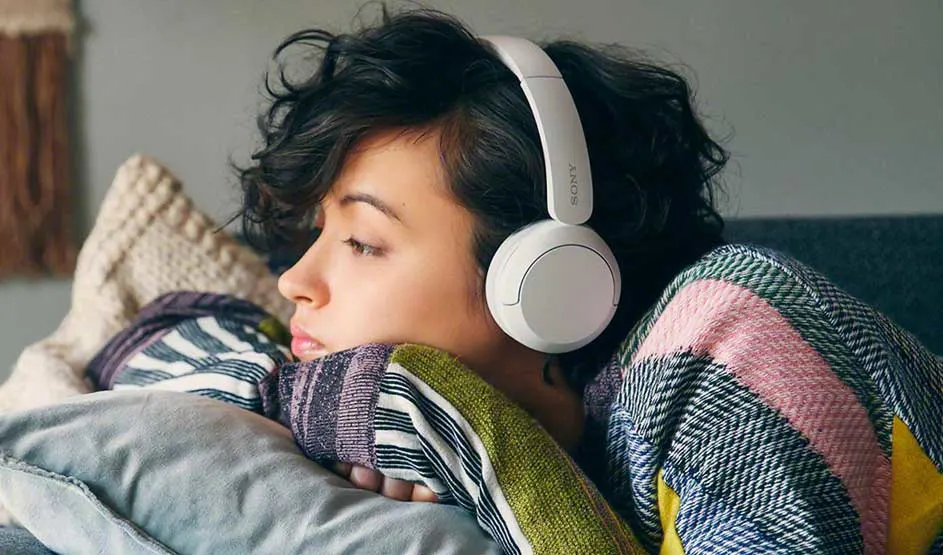 Iemand ligt ontspannen op een bank met een kleurrijke trui aan en draagt witte Sony koptelefoon, omringd door kussens en dekens.