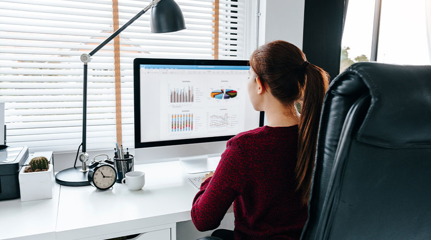 Vrouw zit achter een bureau met een computer waarop grafieken te zien zijn. Er staat een lamp, een klok en een mok.