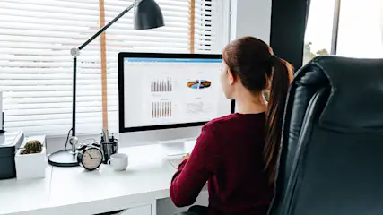 Vrouw zit achter een bureau met een computer waarop grafieken te zien zijn. Er staat een lamp, een klok en een mok.