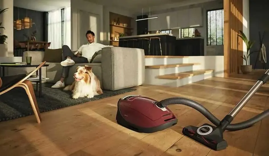 Een rode stofzuiger staat op houten vloer, terwijl een man op de bank ontspant met zijn hond naast zich in een moderne, warme woonkamer.