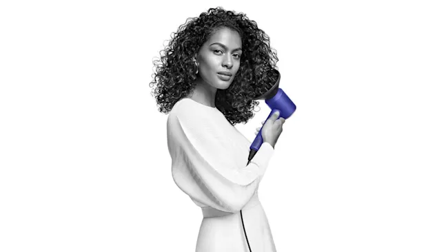 Een vrouw in een witte jurk houdt een blauwe föhn vast tegen een witte achtergrond.