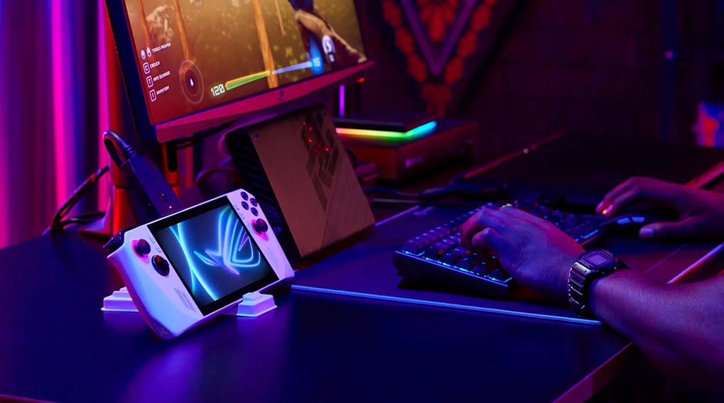 Een gamingopstelling met een handheld console, monitor, toetsenbord, en een persoon die typt.