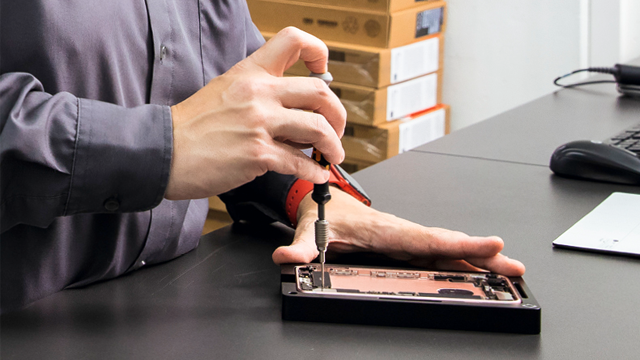Een persoon repareert een zwarte telefoon met een schroevendraaier op een grijs bureau, met dozen en een muis op de achtergrond.