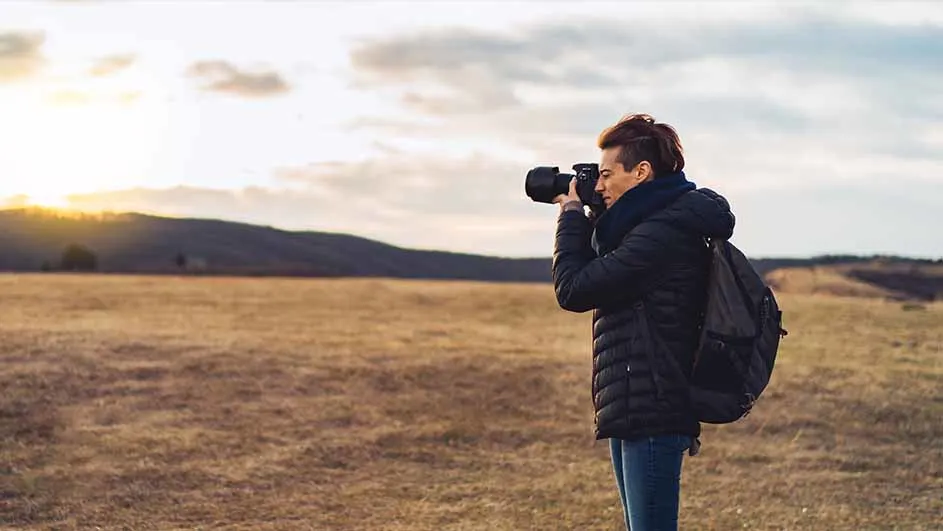 Een fotograaf met een jas en rugzak fotografeert in een veld met heuvels op de achtergrond bij zonsondergang.
