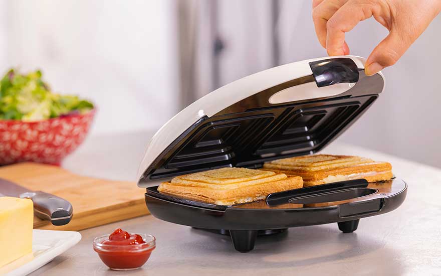 Round sandwich maker on the worktop, two toasted slices inside as a hand lowers the lid, with cheese block and sauce dish nearby.