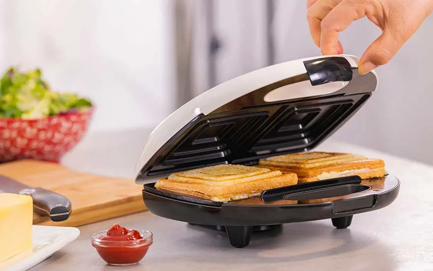 Round sandwich maker on the worktop, two toasted slices inside as a hand lowers the lid, with cheese block and sauce dish nearby.