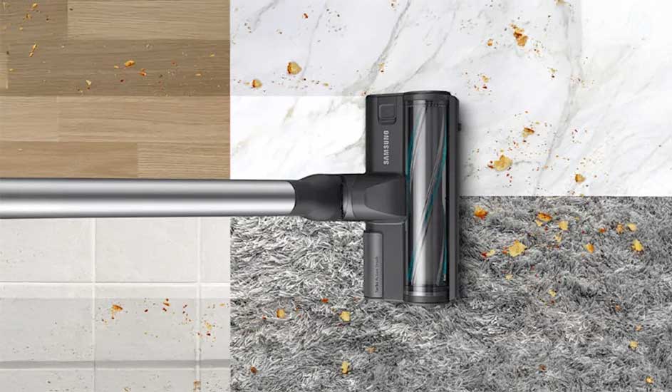Een Samsung stofzuigermond rijdt over vier vloertypen tegelijk—hout, tegels, marmer en een grijs tapijt—en zuigt overal verspreide kruimels op.