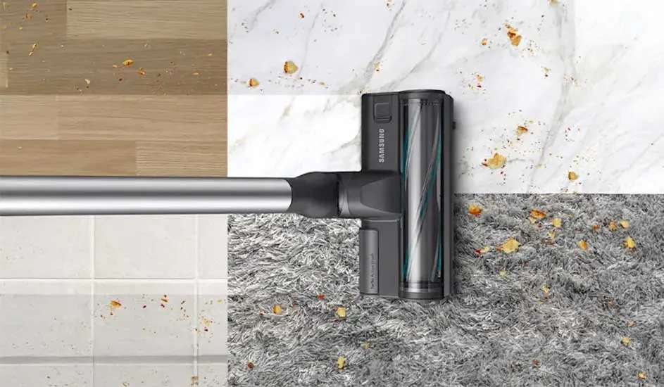 Een Samsung stofzuigermond rijdt over vier vloertypen tegelijk—hout, tegels, marmer en een grijs tapijt—en zuigt overal verspreide kruimels op.