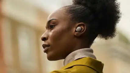 Een vrouw buiten draagt witte draadloze oordopjes van het merk Sony, gekleed in een gele jas en beige coltrui, met haar in een staart.