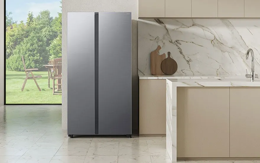 Lichte keuken met grijze Amerikaanse koelkast, marmeren achterwand, beige kasten en grote ramen met tuinzicht buiten.