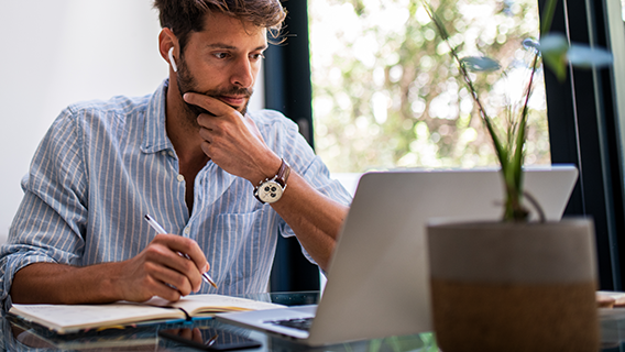 Een man met een gestreept overhemd schrijft in een notitieboek, met een laptop en plant op de achtergrond.