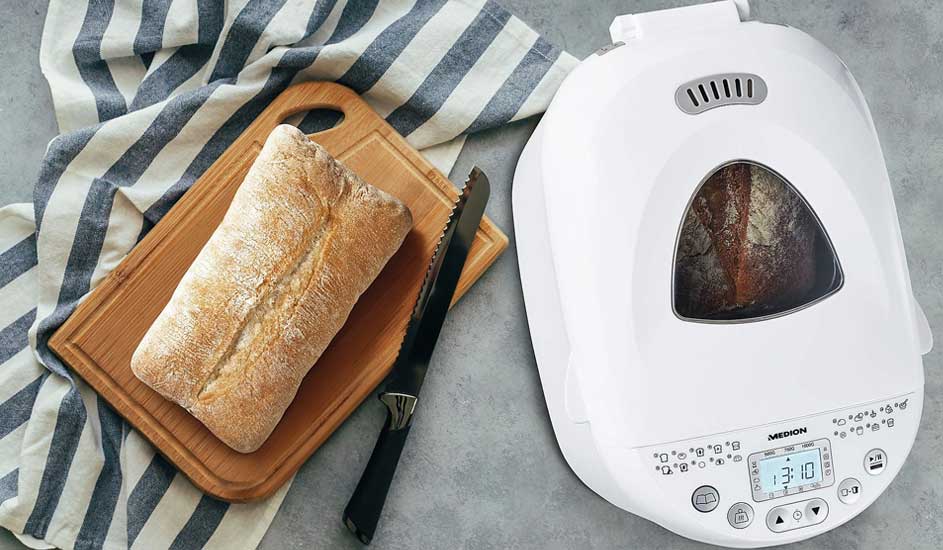 A fresh loaf rests on a wooden board with a bread knife, while another loaf bakes inside the white bread maker beside it.