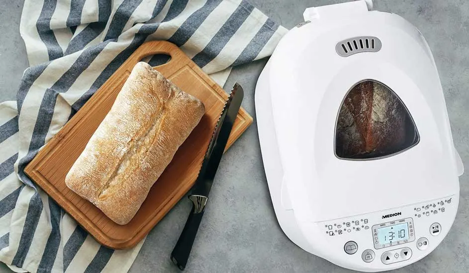 A fresh loaf rests on a wooden board with a bread knife, while another loaf bakes inside the white bread maker beside it.