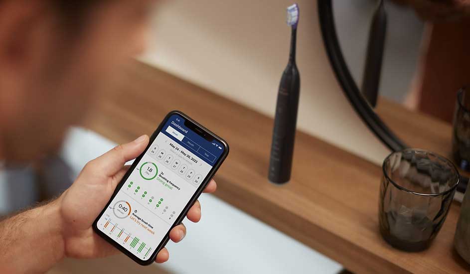 Een hand houdt een smartphone vast met daarop de Oral-B app, naast een zwarte elektrische tandenborstel van het merk Oral-B op de wastafel.