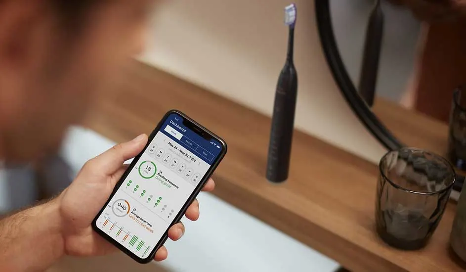 Een hand houdt een smartphone vast met daarop de Oral-B app, naast een zwarte elektrische tandenborstel van het merk Oral-B op de wastafel.