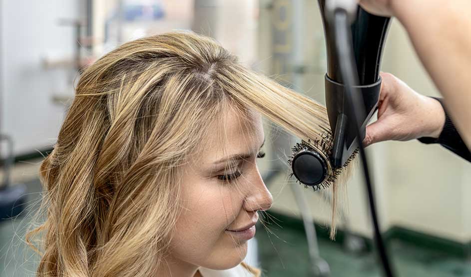 A hairdresser blow-dries and brushes a blonde section smooth, while a woman smiles in a bright salon setting.