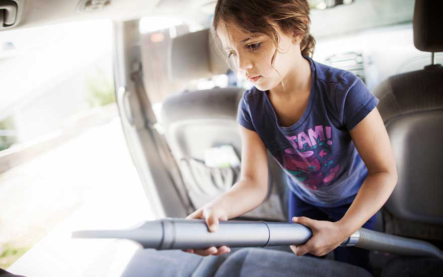 A young girl responsibly vacuums the back seat of a car, focusing intently on removing crumbs and dirt from the upholstery.