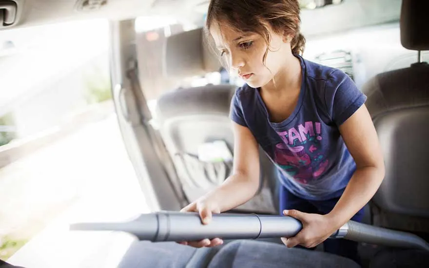 A young girl responsibly vacuums the back seat of a car, focusing intently on removing crumbs and dirt from the upholstery.