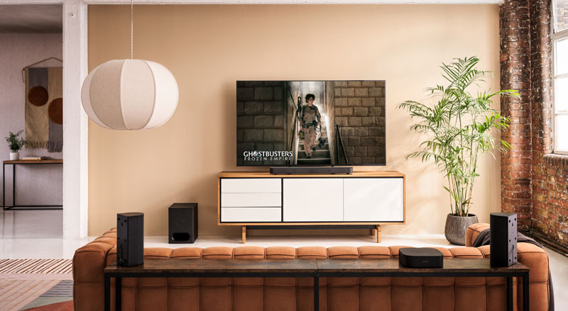 Industriële loftruimte met cognacleren bank, daarachter een wit-houten dressoir met flatscreen-tv die een scène uit Ghostbusters: Frozen Empire afspeelt, en losse surroundspeakers en subwoofer verspreid in de ruimte.