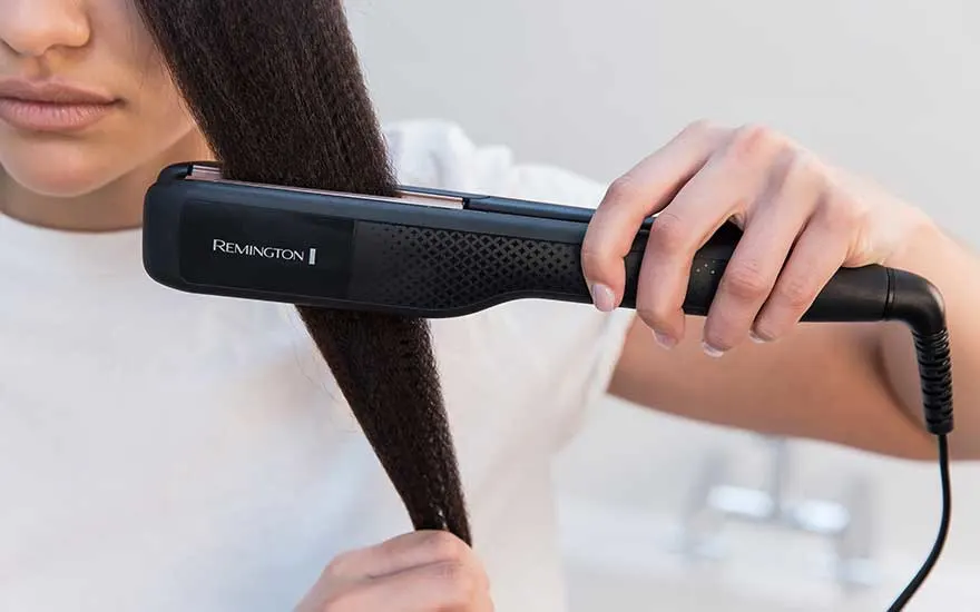 A woman slowly runs a black Remington straightener through a thick section of hair, making it look smooth and sleek.
