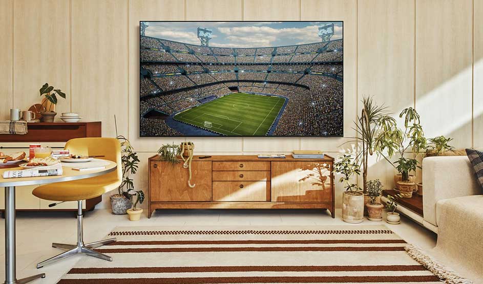 A large TV shows a packed football stadium in a bright, stylish living room with a wooden cabinet, plants, and soft sunlight.