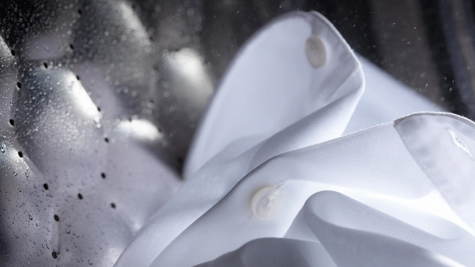 Een witte katoenen blouse met knopen ligt in een zilverkleurige wasmachinetrommel met waterdruppels.