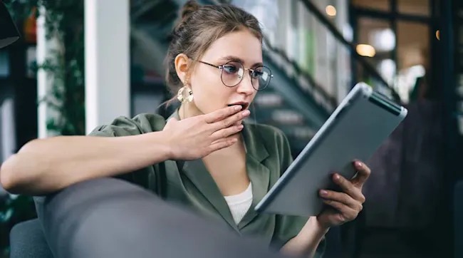 A woman with glasses and earrings looks surprised while holding a tablet in a room with a bookcase in the background.