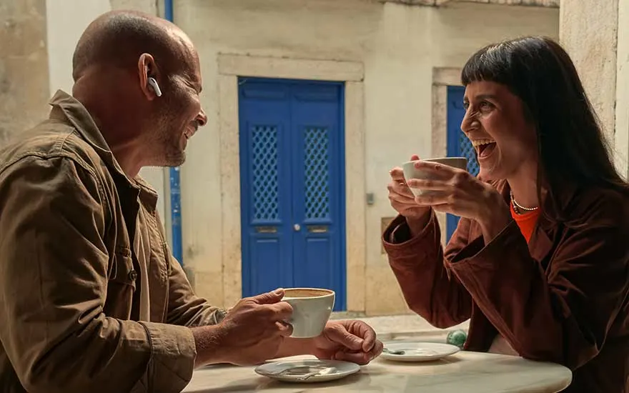 Twee mensen drinken koffie buiten, lachen samen aan een tafel. Er is een blauwe deur op de achtergrond, gezellige sfeer.