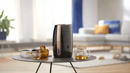 Zwarte cilinder met gouden randen op een tafel met een theepot, kopje, boeken en een plant. Een bank staat op de achtergrond.