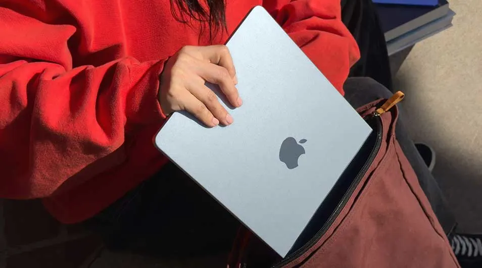 Iemand in rode trui schuift een slanke zilverkleurige laptop veilig in een bruine tas, klaar om onderweg te gaan studeren.