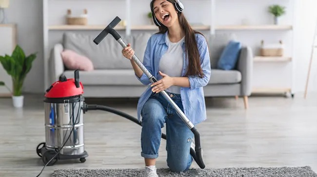 Een vrouw met een koptelefoon knielt op een tapijt en gebruikt een rode en zilveren stofzuiger als gitaar in een woonkamer.