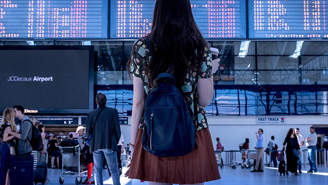 Vrouw met rugzak kijkt naar vertrekborden op een luchthaven, met wachtende mensen op de achtergrond.