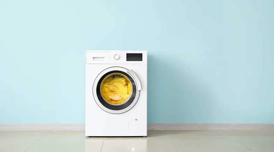 Een witte wasmachine staat tegen een blauwe muur op een witte vloer. De machine bevat gele was.