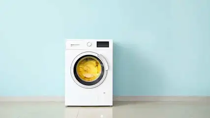 Een witte wasmachine staat tegen een blauwe muur op een witte vloer. De machine bevat gele was.