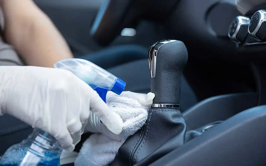 Een persoon met handschoenen sprayt een reinigingsmiddel op een doek en poetst het lederen oppervlak rond de versnellingspook van de auto.