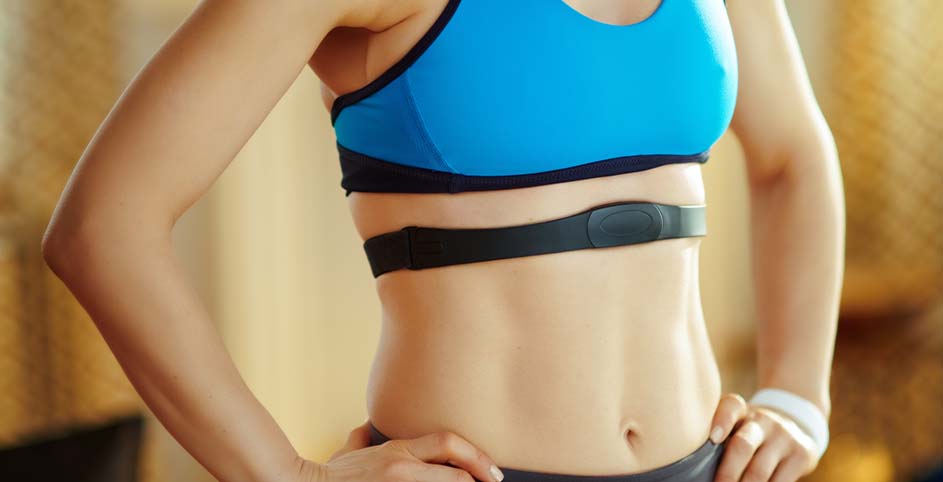 Vrouw in sportkleding met blauwe top en zwarte hartslagmeterband om de borst. Haar handen rusten op haar heupen.