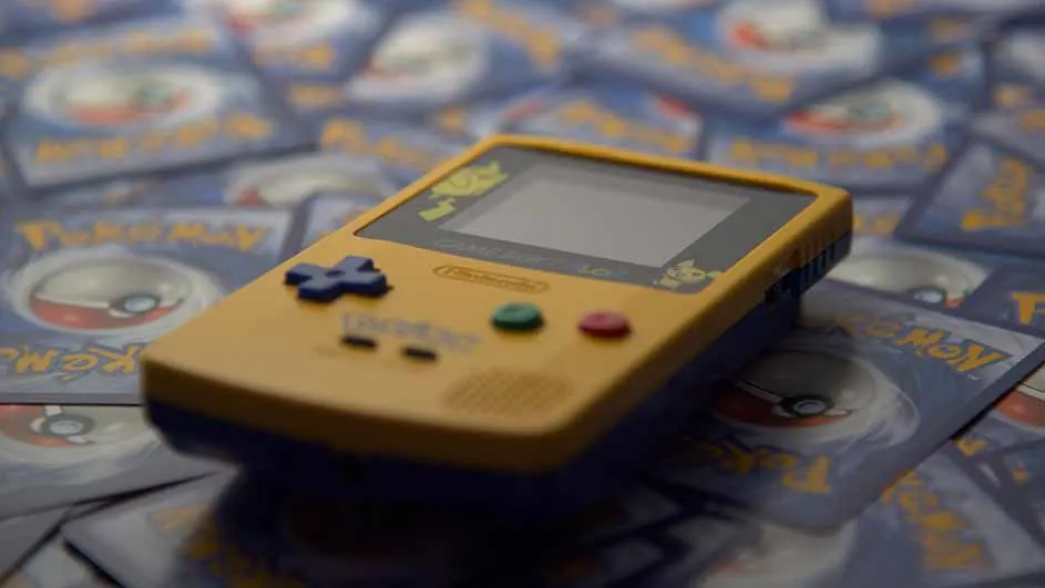 Een gele Game Boy ligt op een stapel Pokémon-kaarten. De Game Boy heeft blauwe, groene en rode knoppen.