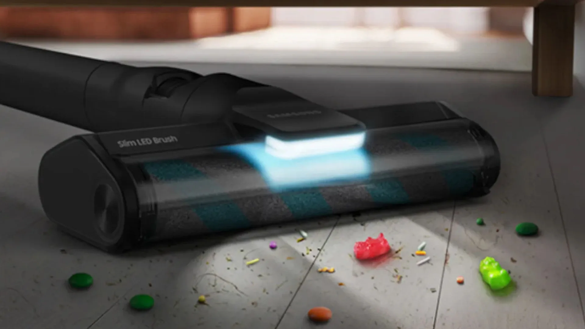 Een zwarte stofzuiger met blauw licht zuigt snoepjes en kruimels op een houten vloer onder een meubelstuk.
