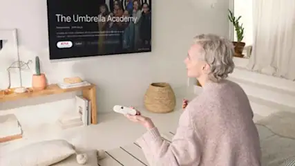 Vrouw kijkt tv met 'The Umbrella Academy'. Ze houdt een witte afstandsbediening vast. Er staat een houten rek met decoraties.
